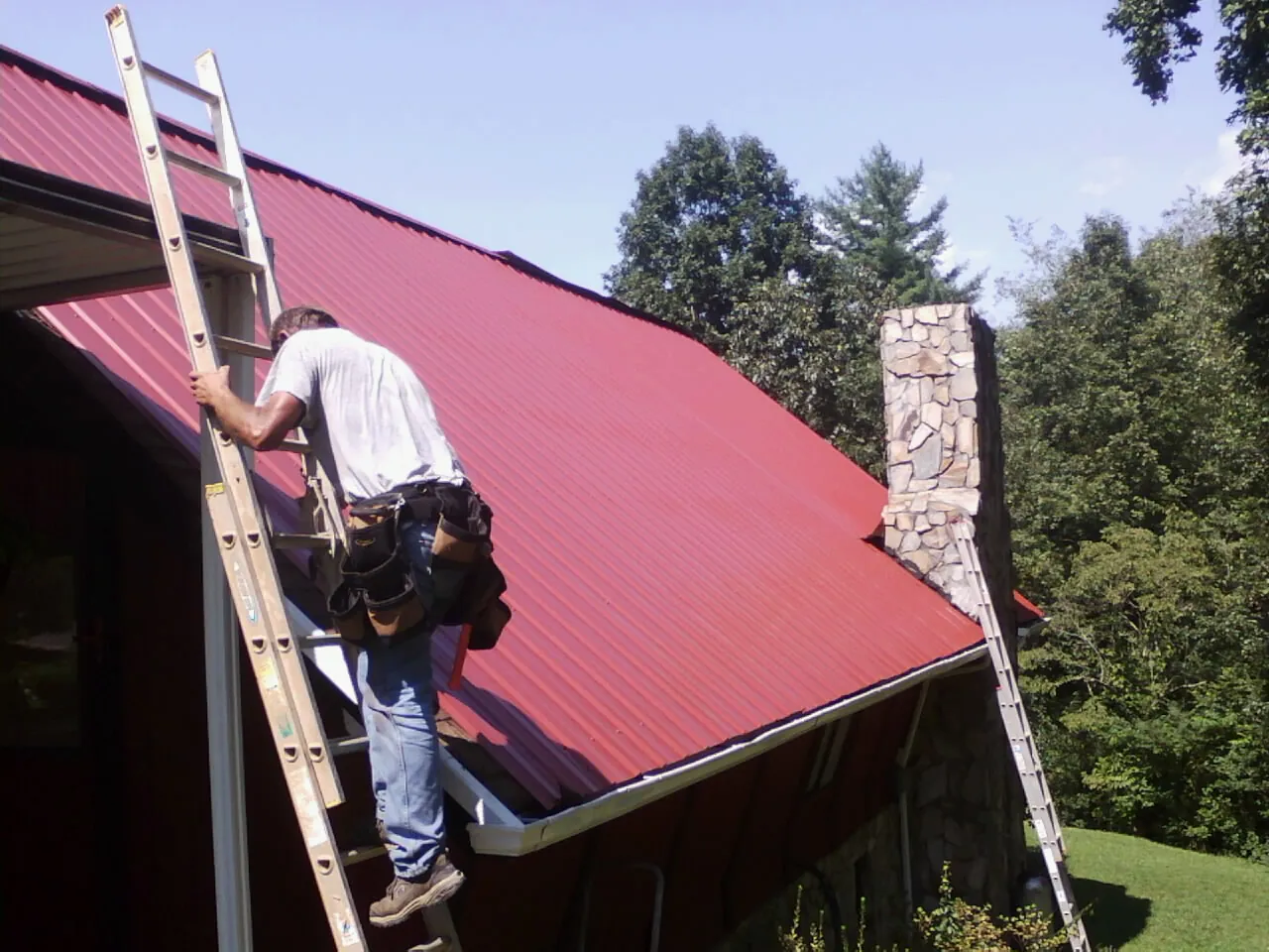 Roofer on a ladder beside a red metal roof and stone chimney — Soffit & Fascia Repair by Roofing Brothers in Voluntown